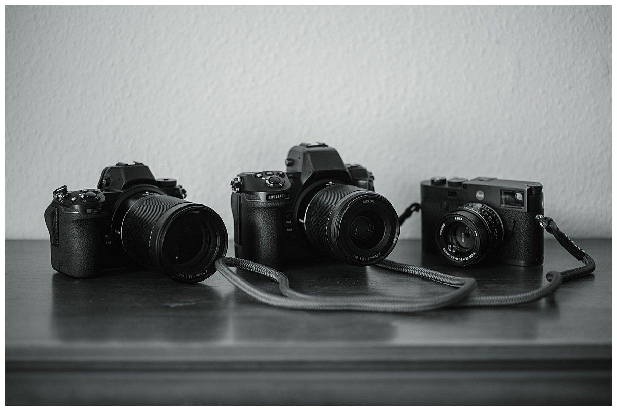 Drei Kameras mit unterschiedlichen Objektivgrößen stehen als Ausrüstung nebeneinander auf einer dunklen Oberfläche vor einer hellen Wand. Das schwarz-weiße Foto zeigt, wie ein Kameragurt locker davor liegt.