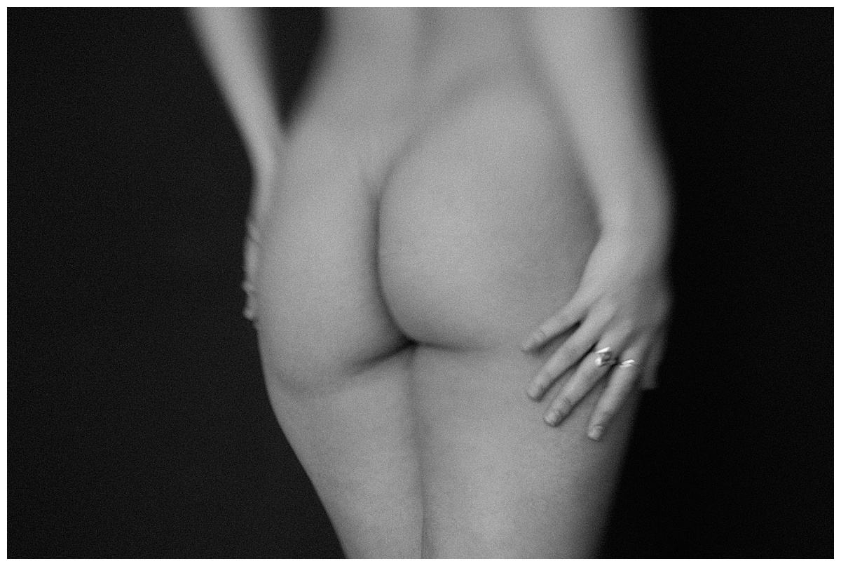 Boudoir_0163 Eine schwarz-weiße, weich fokussierte Aufnahme einer nackten Person von hinten, die eine Hand auf die Hüfte stützt und an deren Finger ein Ring sichtbar ist. Der Hintergrund ist dunkel.