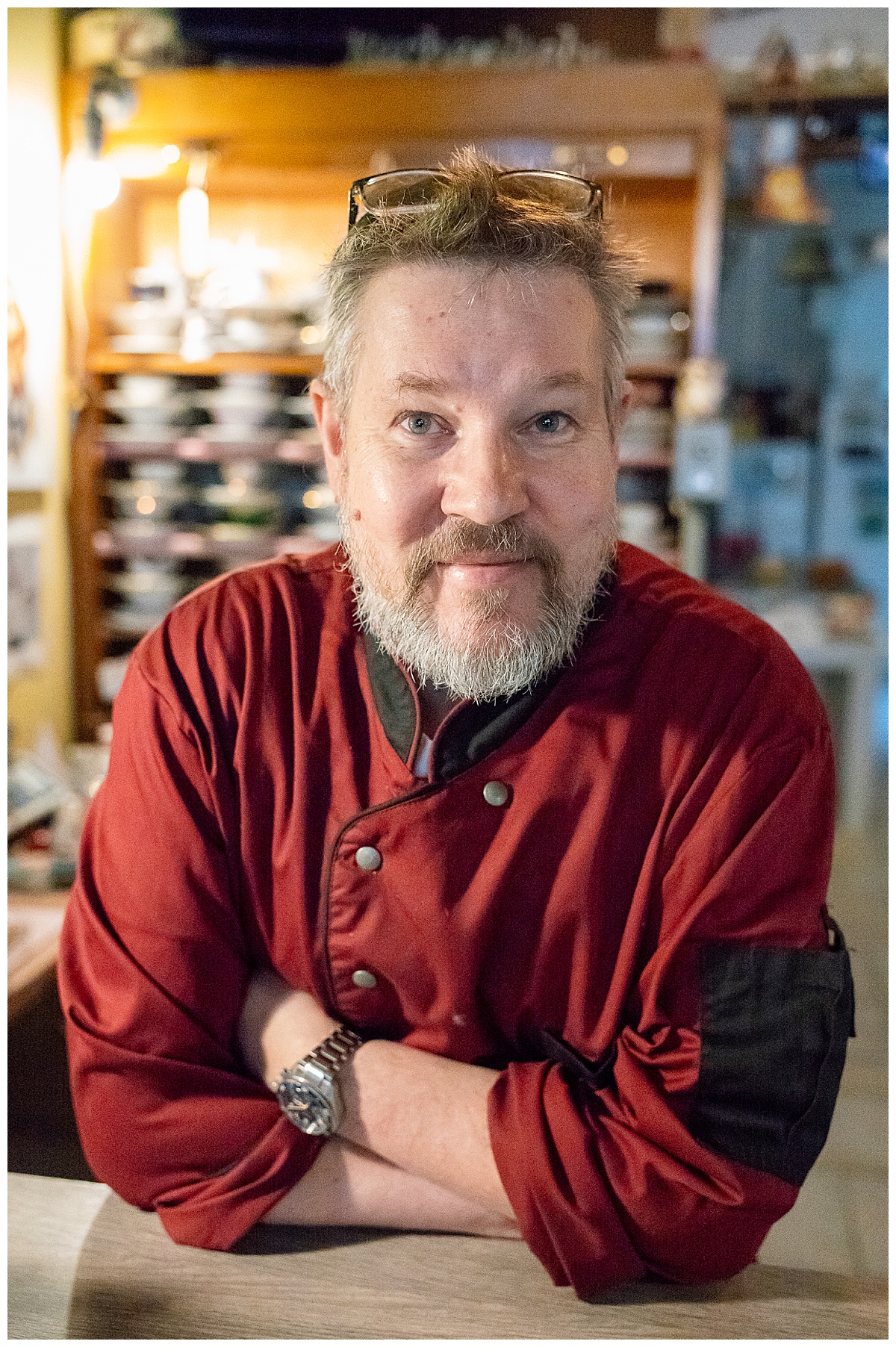 Ein bärtiger Mann in einer roten Kochjacke steht mit verschränkten Armen hinter einem Tresen und lächelt freundlich in die Kamera. Auf seinem Kopf ruht eine Brille, und im schwach beleuchteten Hintergrund sind Regale zu sehen.