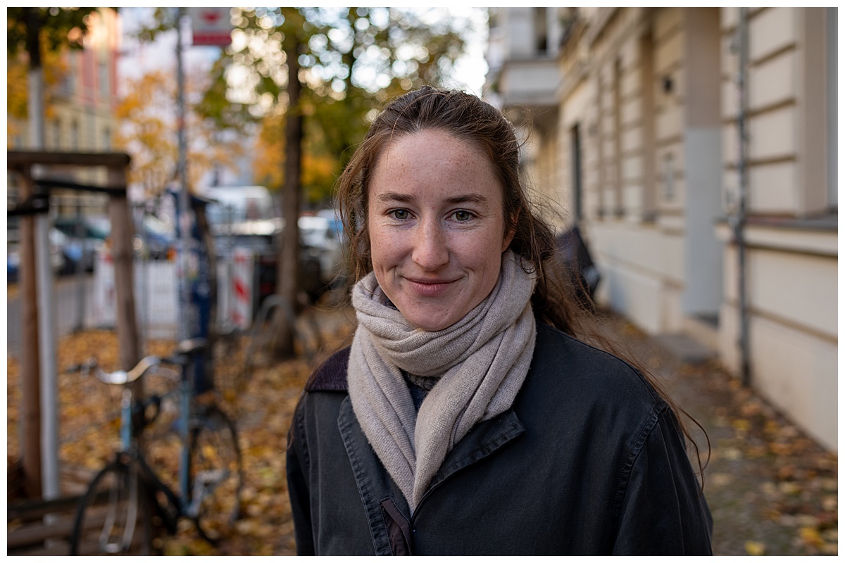 Eine Frau mit langen braunen Haaren, die einen dunklen Mantel und einen hellen Schal trägt, lächelt auf einem mit Herbstblättern und Gebäuden gesäumten Bürgersteig in die Kamera. Im Hintergrund ist ein Fahrrad zu sehen.