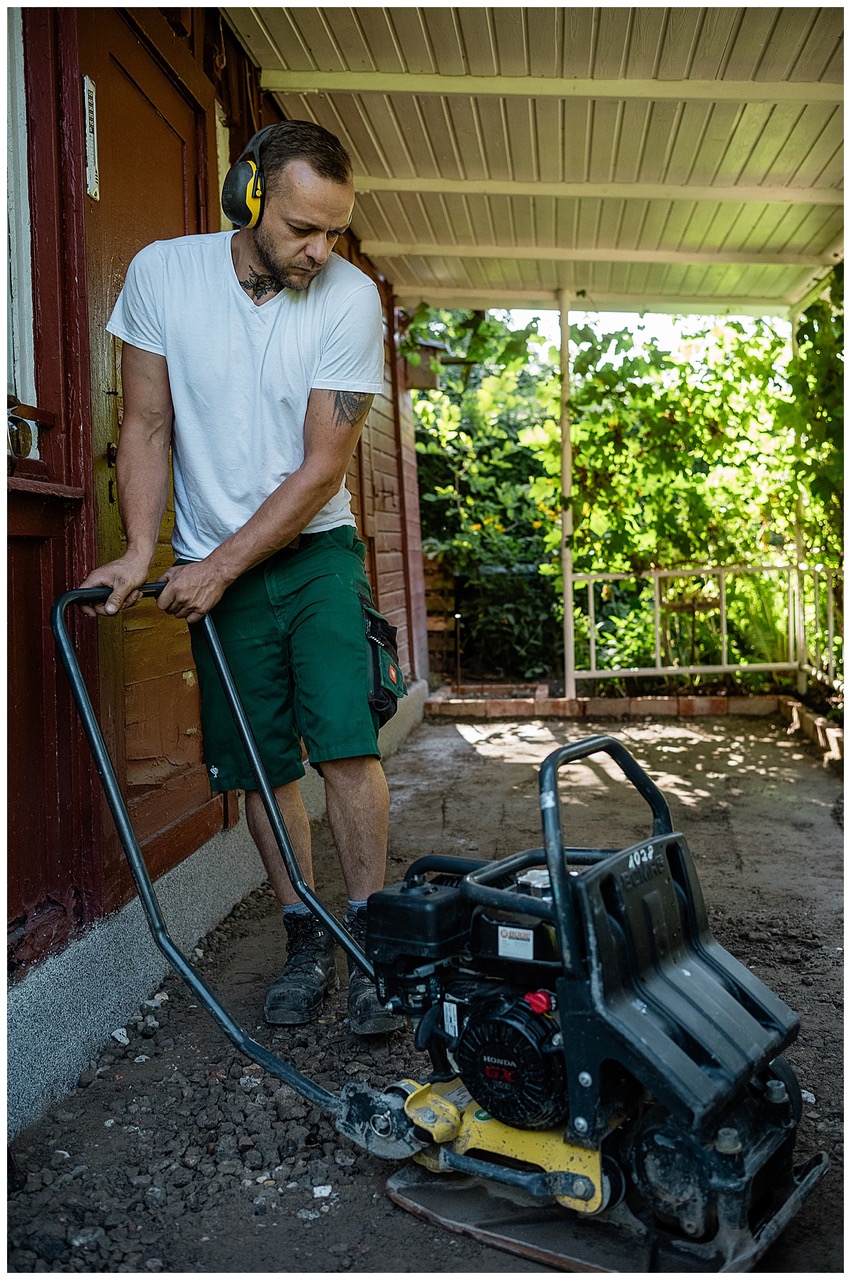 Ein Mann mit Gehörschutz und weißem T-Shirt bedient eine Rüttelplatte auf einer unbefestigten Fläche außerhalb eines Gebäudes unter einer überdachten Veranda mit Grünpflanzen im Hintergrund.