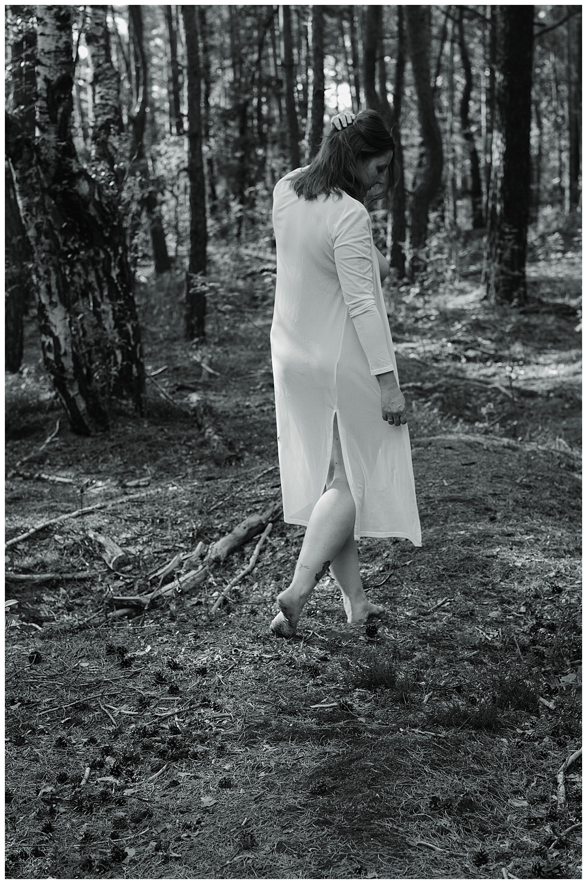 Eine Frau in einem hellen Kleid geht barfuß auf einem Waldweg und schaut mit dem Rücken zur Kamera nach unten. Das Sonnenlicht strömt durch die Bäume und wirft Schatten auf den Boden. Die Szene ist in schwarz-weiß gehalten.